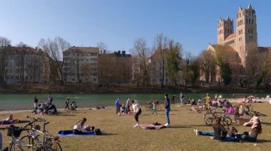 27 Nisan 2022. Münih, Almanya. Münih 'in merkezinde güneşli bir bahar günü. Yerel halk ve turistler, dinlenmek, güneşlenmek ve kentsel doğanın tadını çıkarmak için St. Maximilian Kilisesi yakınlarındaki Isar nehri kıyısında toplandılar.. 