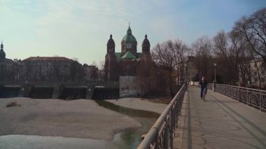 20 Mart 2022. Münih, Almanya. Münih 'in tarihi merkezinde bahar günü Mariannenbruecke ve St. Lukas Kilisesi yakınlarındaki Isar Nehri boyunca berrak mavi gökyüzü altında gezinen yerliler. 