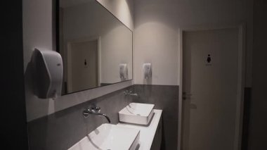 Modern umumi kadınlar tuvaletinde iki beyaz lavabo, duvara monte edilmiş sabun makineleri ve açık bir şekilde işaretlenmiş bir kapı. Parlak iç tasarım, temiz ve işlevsel tasarım ticari kullanım için idealdir. 