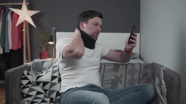 Boyunluk takmış bir adam kanepede oturup video konuşmasında boyun ağrısından yakınıyor. Birine yaralandığını ve çalışamadığını açıklar. Ev ortamı, sıradan sahne. 