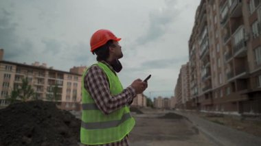 Yansıtıcı yelek ve kareli turuncu gömlekli bir erkek inşaat denetçisi, yüksek katlı bir inşaat alanını incelerken akıllı telefon kullanıyor. Modern mühendislik ve emlak projesi devam ediyor. 