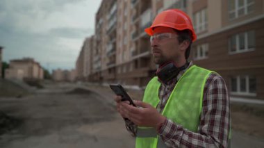 İnşaat müdürü turuncu ekose gömlekli ve güvenli yelekli inşaat alanında telefon kullanıyor. Yüksek katlı konutların gelişimini denetliyor. Teknoloji ve mimari entegrasyonu