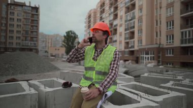 İnşaat mühendisi yüksek binalardaki beton binalarda oturuyor. Kasketli, yansıtıcı yelek giyiyor. Paket kahve içiyor, telefonla konuşuyor ve inşaatı izlerken planları tutuyor.