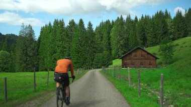 Erkek bir turist, Almanya 'nın Oberstdorf yakınlarındaki Bavyera Alpleri' nde bisiklet yolu boyunca çakıllı bisiklet sürüyor ve gidonu üzerindeki GPS 'ini kontrol ediyor. Etrafı yemyeşil ve ahşap dağ kulübeleriyle çevrili.
