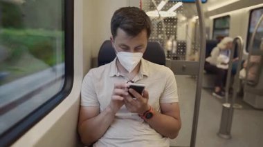 Koruyucu maskeli bir adam, Almanya 'nın Münih şehrinde S-Bahn' de seyahat ederken akıllı telefonunu kullanıyor. Görüntü pandemik dönemdeki toplu taşıma ve dijital izolasyonu yakalıyor