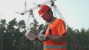 Turuncu üniformalı bir işçi, güvenlik kaskı ormanlık alandaki yüksek voltajlı elektrik hatlarını kontrol ediyor. Pano ve telsizle donatılmış bir şekilde enerji altyapısının güvenliğini ve bakımını sağlıyorlar.. 