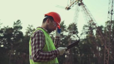 Sert şapka ve güvenlik yeleği giyen elektrik mühendisi, elektrik santralinin yüksek voltajlı elektrik hatlarının yakınında dokümanları olan bir panoyla çalışıyor. Sanayi mühendisi, elektrik hatları ve kuleler. Denetleme
