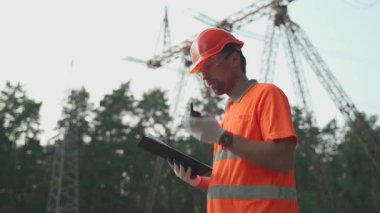 İş kıyafetleri ve turuncu şapkalı bir adam dışarıdaki elektrik hatlarına karşı iş arkadaşlarına bilgi iletmek için telsiz kullanıyor. Radyo ve pano kullanan elektrik santrali çalışanı. 