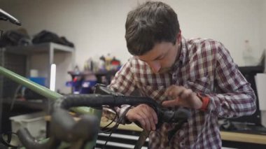 Bisiklet tamircisi tamircisinde çalışıyor, çakıllı bir bisiklete gidon bandı takıyor. Teknisyen bandı ustalıkla gidonun etrafına sarar, atölyede hassasiyet ve kaliteye odaklanır.. 