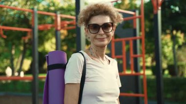 Yaz güneşinde, omuzlarında yoga minderiyle yüzlerinde gülümsemeyle açık hava spor sahasının önünde duran son sınıf kadınının portresi. Açık alanda spor ve eğlence aktiviteleri. Olgun etkinlik. 