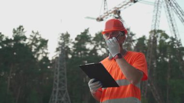 İş kıyafetleri ve turuncu şapkalı bir adam dışarıdaki elektrik hatlarına karşı iş arkadaşlarına bilgi iletmek için telsiz kullanıyor. Radyo ve pano kullanan elektrik santrali çalışanı. 