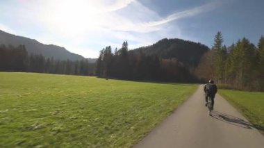 Bisikletçi, Almanya 'nın Bavyera kentinde manzaralı asfalt bisiklet yolu boyunca giderken arkadan görüldü. Çevrelenmiş sonbahar renkleri, bereketli doğa ve dağlar, huzurlu kırsal ve altın güneş ışığı macerayı geliştirir. 