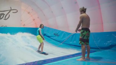Genç, sörf tahtasında dalga simülatöründe kapalı alanda sörf yapıyor. Genç sörfçü, üretilmiş dalgalar üzerine eğitim alırken. Su sporu aktivitesi. Sörfçü çocuk sörf taklitçisi olarak sörf yapmayı seviyor. Sörf eğitimi.