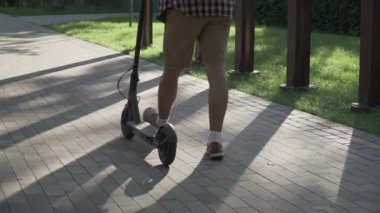 Ekose gömlekli, güneş gözlüklü, güneşli bir parkta, yaz havasında elektrikli scooter süren beyaz bir adam. Konu ekolojisi, çevre koruma, araba egzoz gazlarından kaynaklanan hava kirliliği. Eko taşımacılığı.