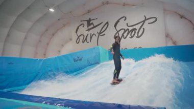 Genç, sörf tahtasında dalga simülatöründe kapalı alanda sörf yapıyor. Genç sörfçü, üretilmiş dalgalar üzerine eğitim alırken. Su sporu aktivitesi. Sörfçü çocuk sörf taklitçisi olarak sörf yapmayı seviyor. Sörf eğitimi.