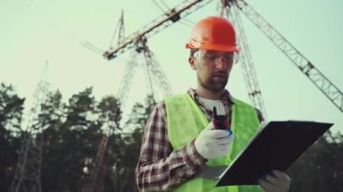 Yüksek voltaj kulesinin yanında telsiz ve pano kullanarak elektrikli ekipman işçisi. Sanayi mühendisi, elektrik hatları ve kuleler. Dışarıdaki şanzıman hattının yakınındaki elektriklendirmeyi denetliyorum..