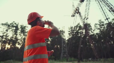 Öğle yemeği molasında inşaat işçisi. Teknisyen, kasklı bir mühendis öğle yemeği yiyor ve işyerindeki elektrik hattı altyapısıyla kahve içiyor. Elektrikçi abur cubur yer ve kahve içer..