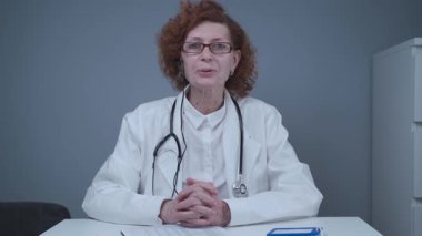 Kulaklık takmış yaşlı kadın pratisyen kamerayla görüntülü görüşme yapıyor, hastaya görüntülü konuşma yoluyla danışmanlık yapıyor. Doktor görüntülü görüşme için danışmanlık yapıyor. Web kamerasından görüntüle.