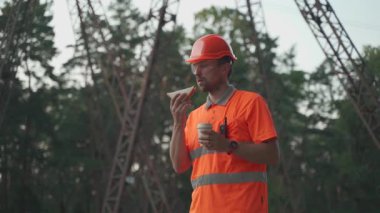 Engineer having lunch near power lines and electricity pylons in the field. An electrical substation worker in a protective helmet eats a sandwich and drinks coffee during a break at the workplace.