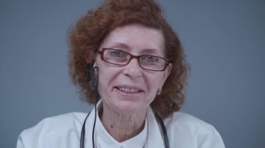 Beyaz laboratuvar önlüğü ve kulaklık takmış yaşlı kadın pratisyen kameraya bakan video iletişimi uzmanı. Webcam görüntüsü. Hastane ofisinden video konferansı..