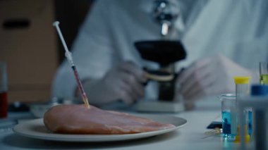 Gıda kalite kontrol uzmanı laboratuvarda mikroskopla çalışıyor. Çiğ ete antibiyotik enjekte edilen şırıngalar, tavukta genetiği değiştirilmiş organizmalar, ilaçlarda ve yiyeceklerde kimyasallar.