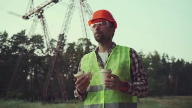 Hungry worker in overalls in electric power business eats sandwich and drinks takeaway coffee at workplace near power lines. Power worker eating snack with hot drink near high voltage power line.