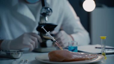 Gıda kalite kontrol uzmanı laboratuvarda mikroskopla çalışıyor. Çiğ ete antibiyotik enjekte edilen şırıngalar, tavukta genetiği değiştirilmiş organizmalar, ilaçlarda ve yiyeceklerde kimyasallar.