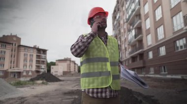 Koruyucu iş kıyafetleri içindeki inşaatçı ellerinde eşyalar olan evlerin inşaatında telefonla konuşuyor. Projede yürürken inşaat sürecini Foreman kontrol ediyor.