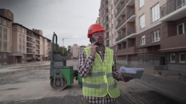 İnşaat alanında çalışan ustabaşı inşaat planı tutuyor ve inşaat alanında cep telefonuyla konuşuyor. Kasketli, yelekli, yürüyen ve telefonda konuşan biri..