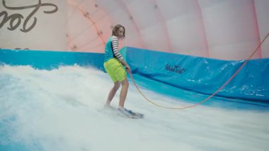 Su sporu aktivitesi, denge konsepti. Küçük sporcu ev içinde sörf yapmaktan hoşlanıyor. Gençler spor kompleksinde simülatörde dalgalarda sörf yapıyor. Sörf koçu ve öğrenci dalga simülatöründe toplandı.