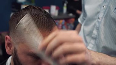 Bir berber dükkanında, profesyonel bir berber saç kesimi sırasında ıslak saçlarını ustalıkla tarar. Her kesin hareket, erkek bakımındaki beceriyi, stili ve dikkati yansıtır.. 