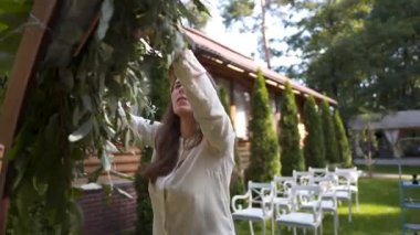 Profesyonel çiçekçi düğün kaydı için festival kemerini dekore ediyor. Kadın düğün dekoru kuruyor. Dekoratör, açık hava düğün törenini çiçekler ve yeşilliklerle süslüyor..