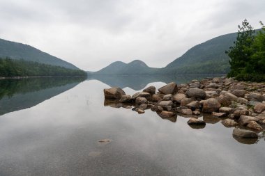 Acadia Ulusal Parkı 'ndaki Ürdün Pond' un sakin, yansıtıcı suları, ikonik Bubble Dağları, kayalık bir kıyı şeridi ve sakin, bulutlu gökyüzünün altındaki yemyeşil ormanları gösteren huzurlu bir manzarası..