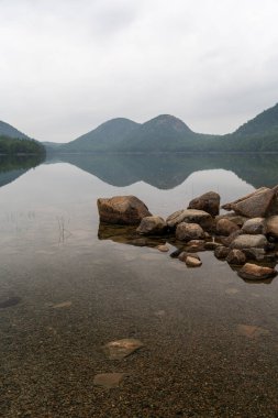 Acadia Ulusal Parkı 'ndaki Ürdün Pond' un sakin, yansıtıcı suları, ikonik Bubble Dağları, kayalık bir kıyı şeridi ve sakin, bulutlu gökyüzünün altındaki yemyeşil ormanları gösteren huzurlu bir manzarası..