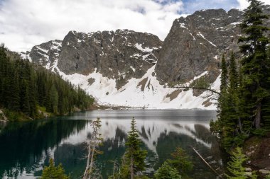 Blue Lake near North Cascades National Park and Okanogan-Wenatchee National Forest, with its crystal-clear waters reflecting snow-capped peaks, lush evergreens, and a dramatic, cloudy sky. clipart