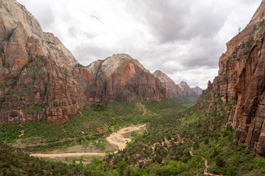 Zion Ulusal Parkları 'nın kırmızı kaya kanyonunun çarpıcı manzarası, bereketli bir vadide dramatik uçurumlar yükseliyor. Sarmal nehir ve canlı yeşillik kontrastı yukarıdaki karamsar, bulutlarla dolu gökyüzüne karşı.