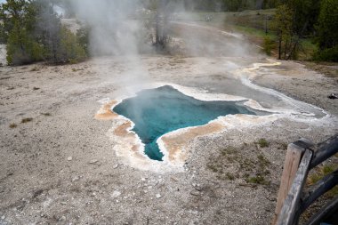 A vibrant hot spring in Upper Geyser Basin, Yellowstone National Park. The colorful geothermal feature is surrounded by rugged terrain and clear blue skies, highlighting Yellowstones natural wonder. clipart