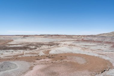 Arizona 'daki Petrified Forest Ulusal Parkı' nın kurak ve aşınmış manzarası. Katmanlı tepeler, kurak akıntılar ve çöl arazisi boyunca uzanan açık mavi bir gökyüzü..