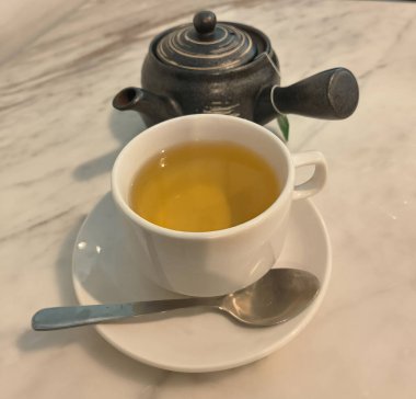 Beyaz porselen fincanın üzerinde sarmal kapaklı koyu kil Japon çaydanlığı altın-yeşil çayla dolu. Çaydanlığın yan sapı ve çıkıntısı var. Mavi kaşıklı fincan üstü fincan. Geleneksel ve modern çay seti.