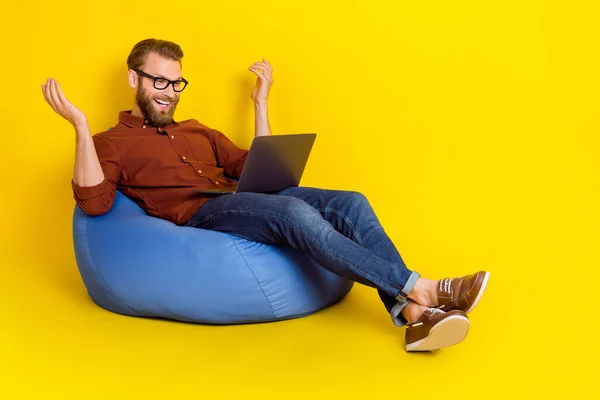 Full length photo of positive man sit comfy bag speak video call netbook isolated on yellow color background.