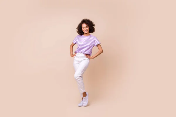 楽観的な可愛い女の子の完全な長さの写真は ストライプTシャツの白いズボンの靴腕上の腰に隔離されたベージュ色の背景 — ストック写真