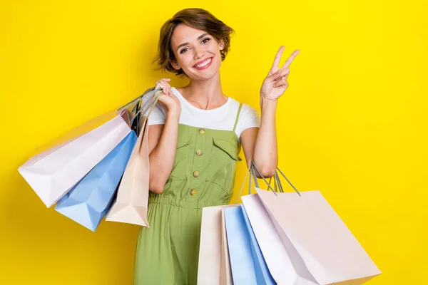 年轻积极友善的女孩购物狂的照片 展示V字形符号 顾客拿着包黄色背景的新衣服销售 — 图库照片