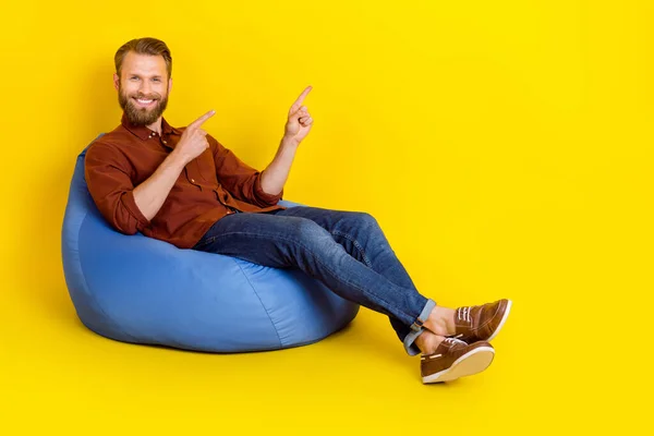 Full body portrait of positive guy sit bag direct fingers empty space isolated on yellow color background.