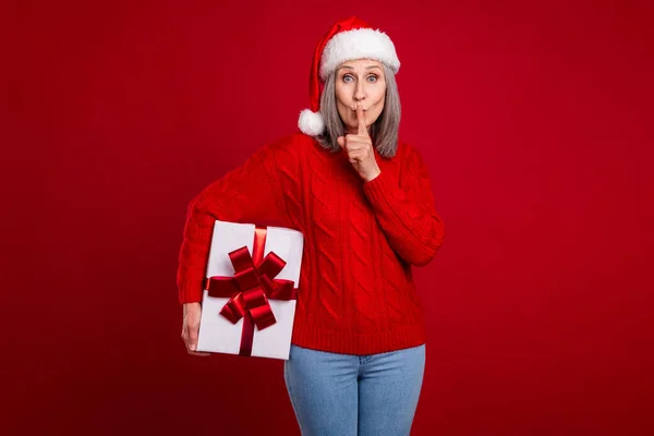 Photo of funny cute secretive granny hold giftbox make shh sign wear hat pullover isolated red color background.