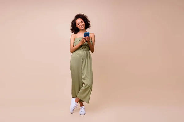 stock image Full size photo of good mood girl with wavy hairstyle dressed khaki overall sneakers writing post isolated on beige color background.