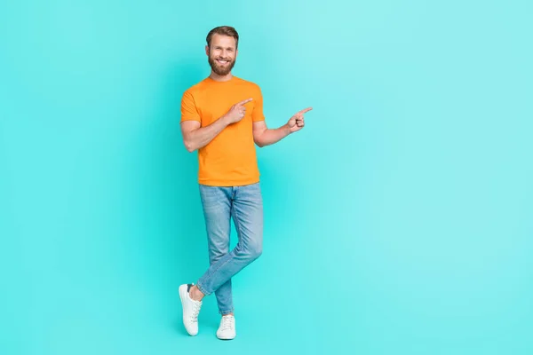 フルサイズの写真の素敵なひげ男インデックス空のスペース着用オレンジTシャツジーンズスニーカー隔離された上のティール色背景 — ストック写真
