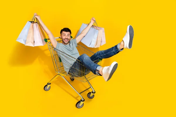 喜びに満ちた人の完全な長さの写真座っている市場トロリーホールドショッピングバッグ黄色の色の背景に隔離 — ストック写真