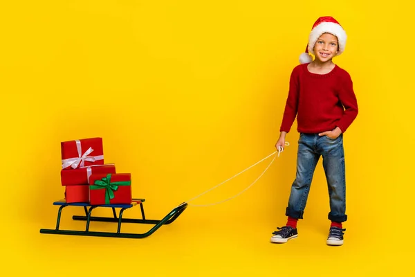 フルサイズの写真の魅力的な小さな男の子プルサンタクラスのそりプレゼントヘルパー着用トレンディーな赤い服黄色の背景に隔離 — ストック写真