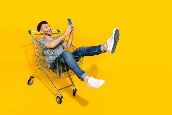英俊快乐男人坐在商场推车上的全副照片用黄色背景隔离电话 — 图库照片