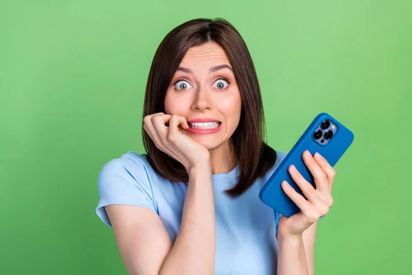 Portrait of nervous pretty person hold telephone biting finger nail isolated on green color background.